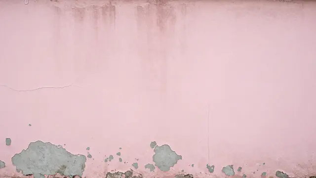 A weathered pink plaster wall with peeling paint revealing gray cement and fine cracks