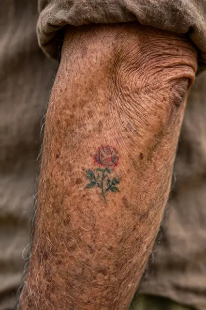 Close-up of an older man's forearm showing a small faded traditional rose tattoo