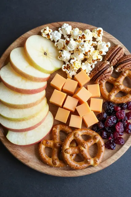 Overhead view of a round wooden board arranged with apple slices, cheese, popcorn, pretzels, pecans and dried cranberries.