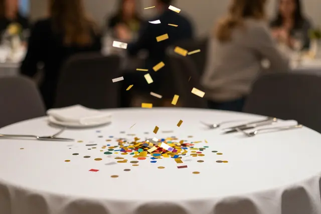 Metallic and colorful confetti falls onto a white-clothed round event table with blurred seated guests in the background
