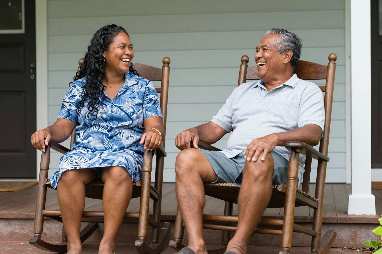 Two Pacific Islander people laugh together while seated in wooden rocking chairs on a residential front porch.