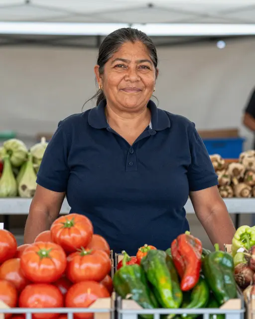 Mature Latina vendor stands behind crates of fresh tomatoes and peppers at a market stall, smiling at the camera