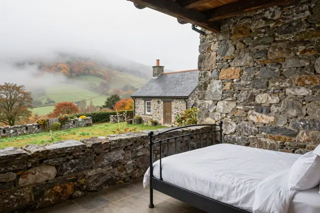 Iron bed in a stone walled room overlooking misty autumn hills and a cottage