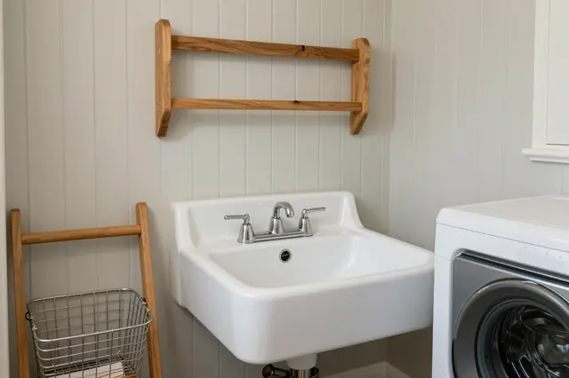 A tidy farmhouse laundry nook features a white apron sink, wooden drying rack and wire basket next to a washing machine.