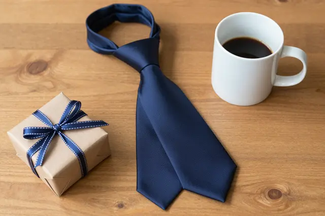 A navy silk tie, white coffee mug, and small wrapped gift sit on a light wooden table.
