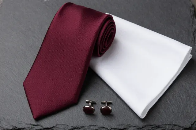 Rolled burgundy tie, white pocket square and matching cufflinks rest on dark slate stone