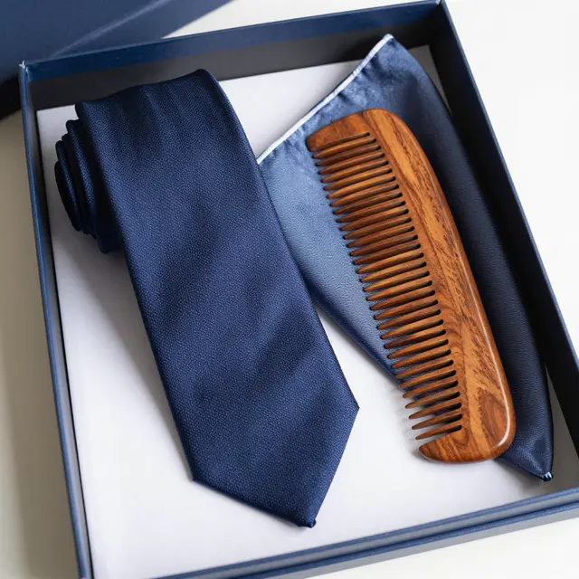 Top view of an open navy gift box holding a rolled silk tie, pocket square and wooden comb on a white surface