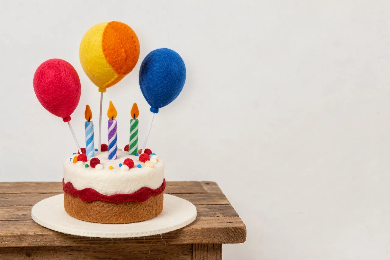Handmade felt birthday cake with candles and balloons sits on a wooden table with empty right-side copy space