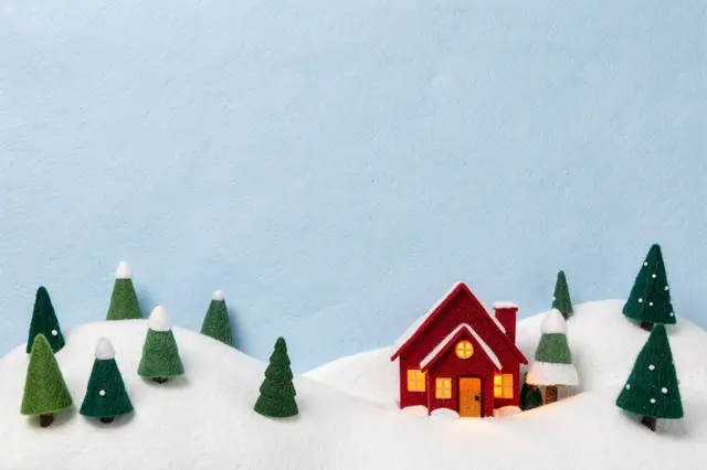 Felt snowy hills with tiny pine trees and a lit red cottage sit under a pale blue empty sky