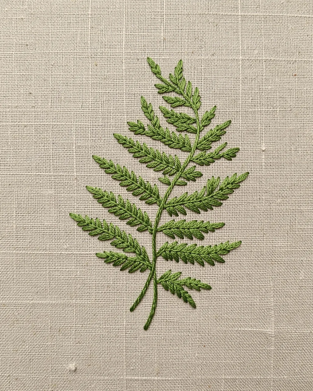 Fine green embroidery stitches form a fern frond against a natural woven linen background.