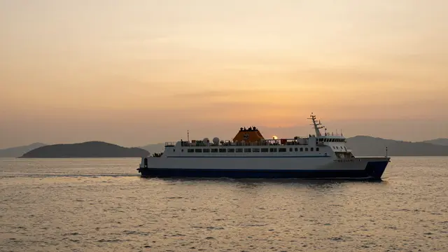 A passenger ferry sails across calm golden water with distant islands under a warm hazy sunset sky.