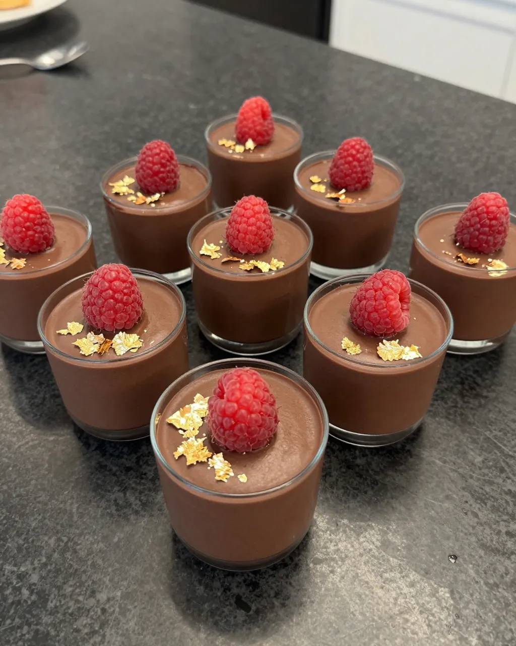 Arranged glass chocolate mousse cups topped with raspberries and gold leaf on a dark marble countertop