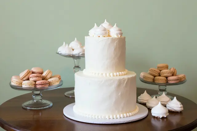 Two tier white cake with meringue accents sits alongside macarons and meringues on glass stands on a wooden table against