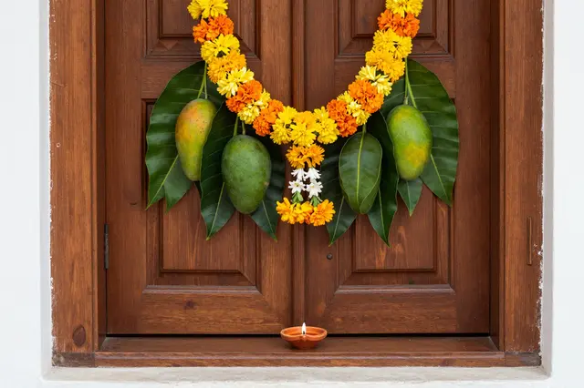 Marigold and mango leaf garland hangs over a carved wooden door with a lit diya on the threshold