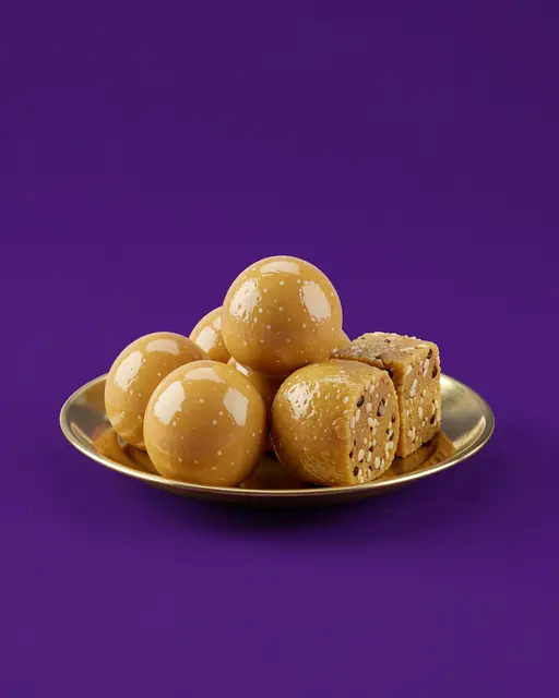 Glossy ladoos and barfi style sweets rest on a small brass plate against a solid deep purple background