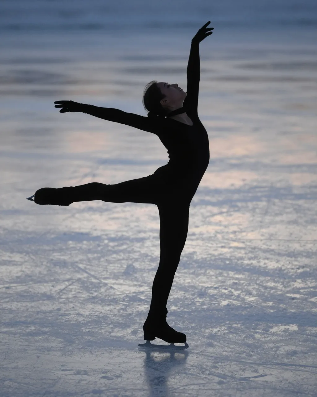 A figure skater holds a graceful ballet pose on smooth frozen ice at dusk