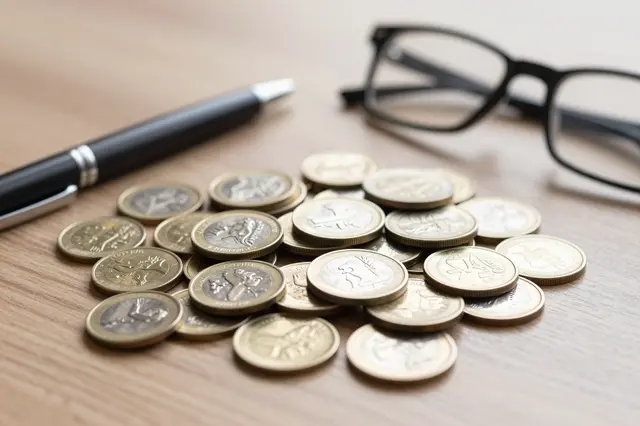 Pile of bimetal coins, a black pen, and eyeglasses rest on a light wooden desk surface.