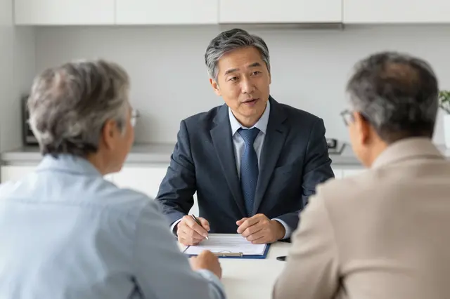 A financial advisor speaks with two senior people seated across a kitchen table, viewed from behind the couple.
