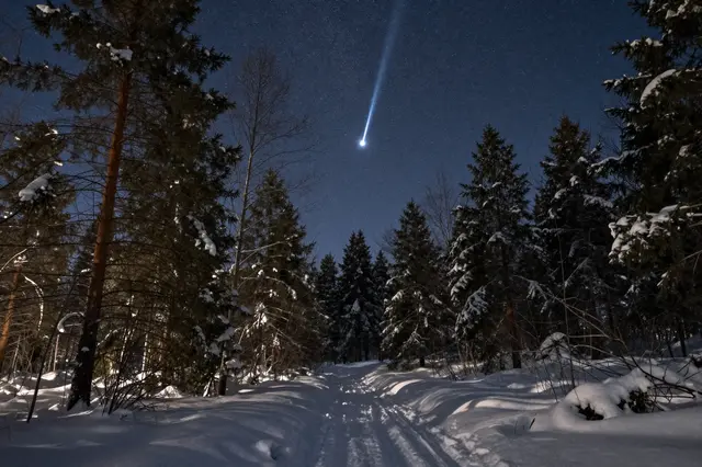 A bright streaking fireball meteor hangs in the night sky above a snow covered winter forest trail.