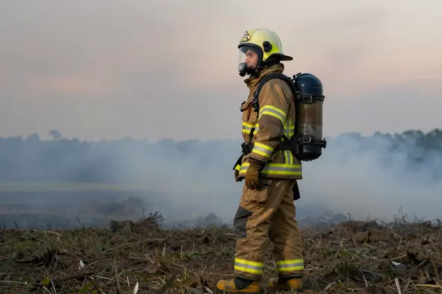 A side view of a firefighter in full protective gear standing beside a smoky field at dusk