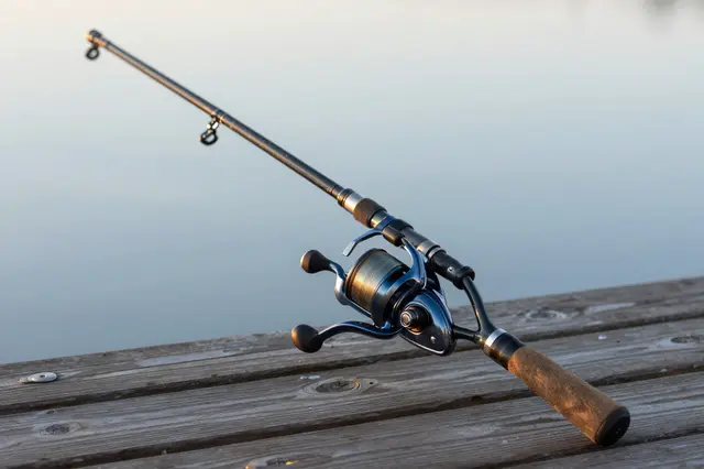 A spinning fishing rod rests diagonally on a weathered wooden dock beside calm water at dawn