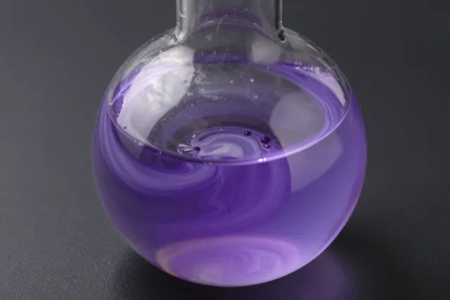A close-up view of an unmarked round glass flask holding swirling purple liquid against a dark background