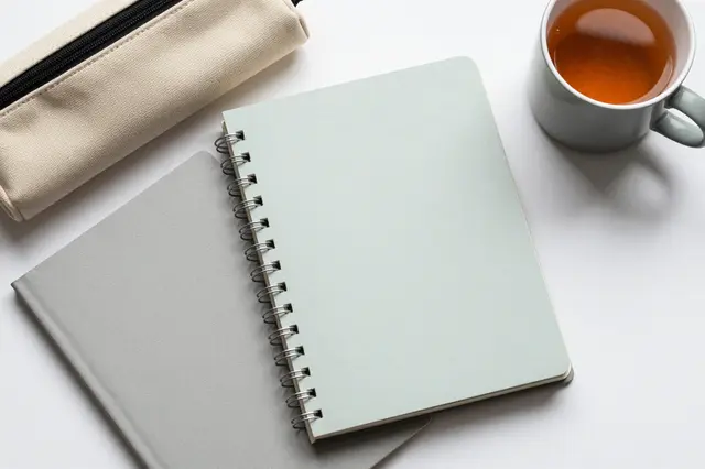 Top-down flat lay of muted notebooks, a canvas pencil case, and a mug of tea on a white surface