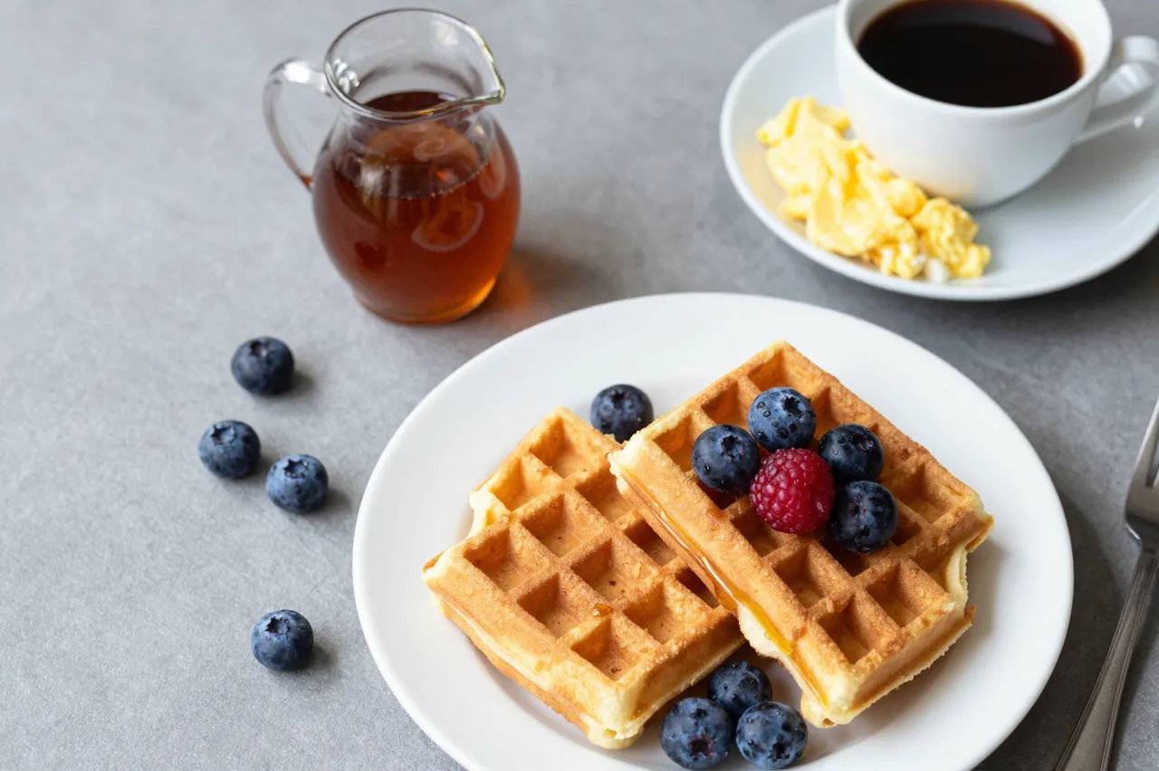 Top-down flat lay of waffles, berries, maple syrup, scrambled eggs and coffee arranged on a gray tabletop.