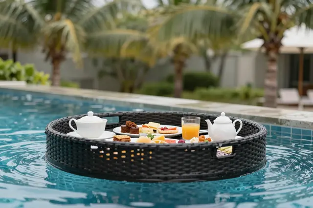 A woven floating breakfast tray holding food and drink rests on turquoise resort pool water with palm trees in the