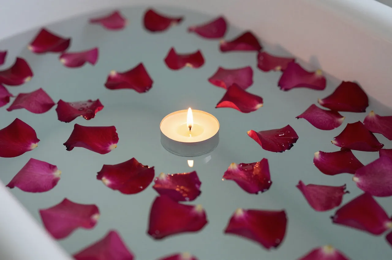 A lit floating tealight candle sits surrounded by red rose petals on the surface of bath water in a white tub