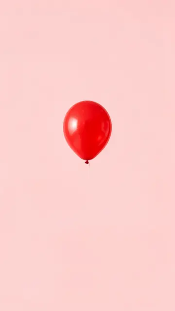 A single glossy red balloon floats centered against a solid pale pink background with empty surrounding space.