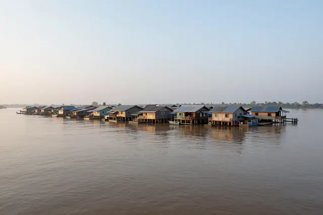 A line of stilted wooden floating houses stretches across a wide brown misty river at early dawn