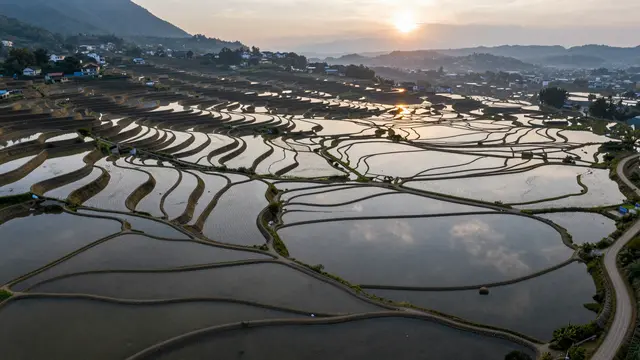 Aerial view of water-filled rice terraces reflecting the sky at sunrise with distant mountain and village views