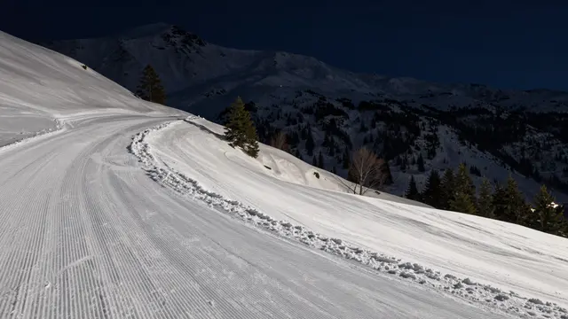 A curved groomed snowy ski run stretches up a moonlit mountain slope under a dark night sky.