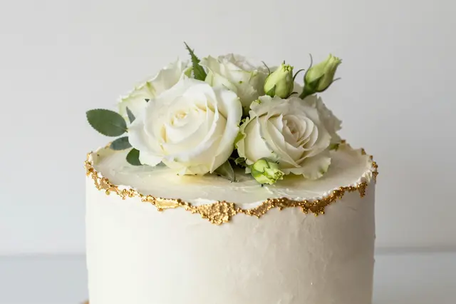 A single tier white frosted cake topped with fresh white roses, green leaves and textured gold edge trim.