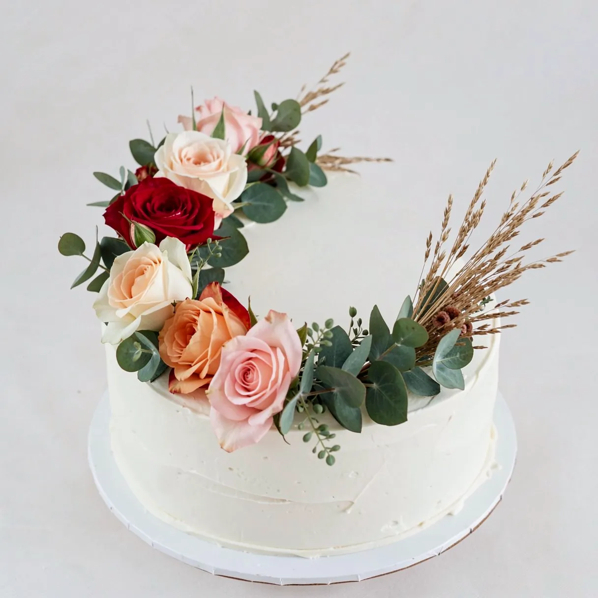A white buttercream cake decorated with a crescent of fresh roses, eucalyptus, and dried grasses on a cake stand.