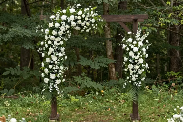 A wooden wedding arch decorated with white flowers and green foliage stands in a sun dappled forest clearing.