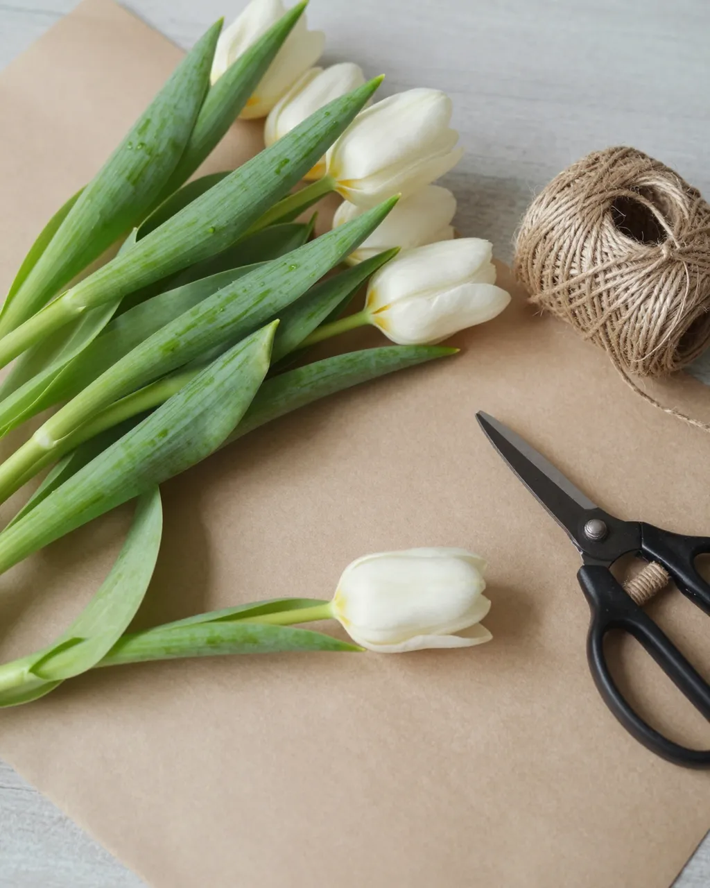 Flat lay arrangement of white tulips, kraft paper, jute twine and pruning shears on a light wood table in soft daylight