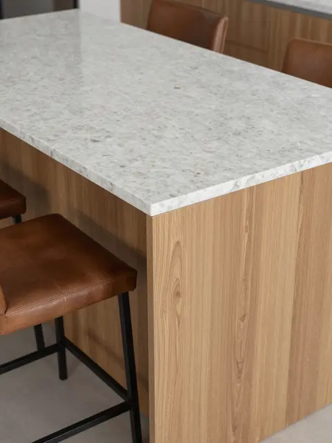 Partial view of a fluted oak kitchen island with a light quartzite top and brown leather stools.