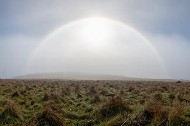 A pale fogbow arcs across a misty sky above a grassy moorland