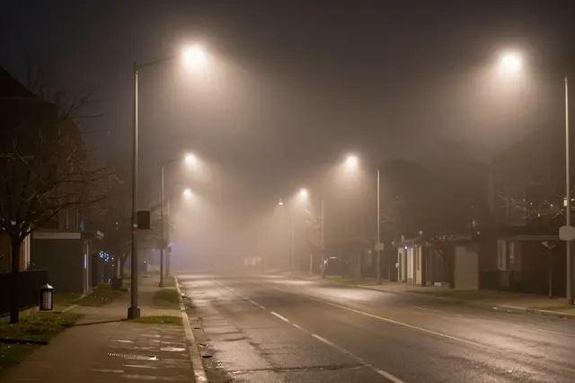 An empty wet city street stretches into thick fog, lit by glowing streetlights at night