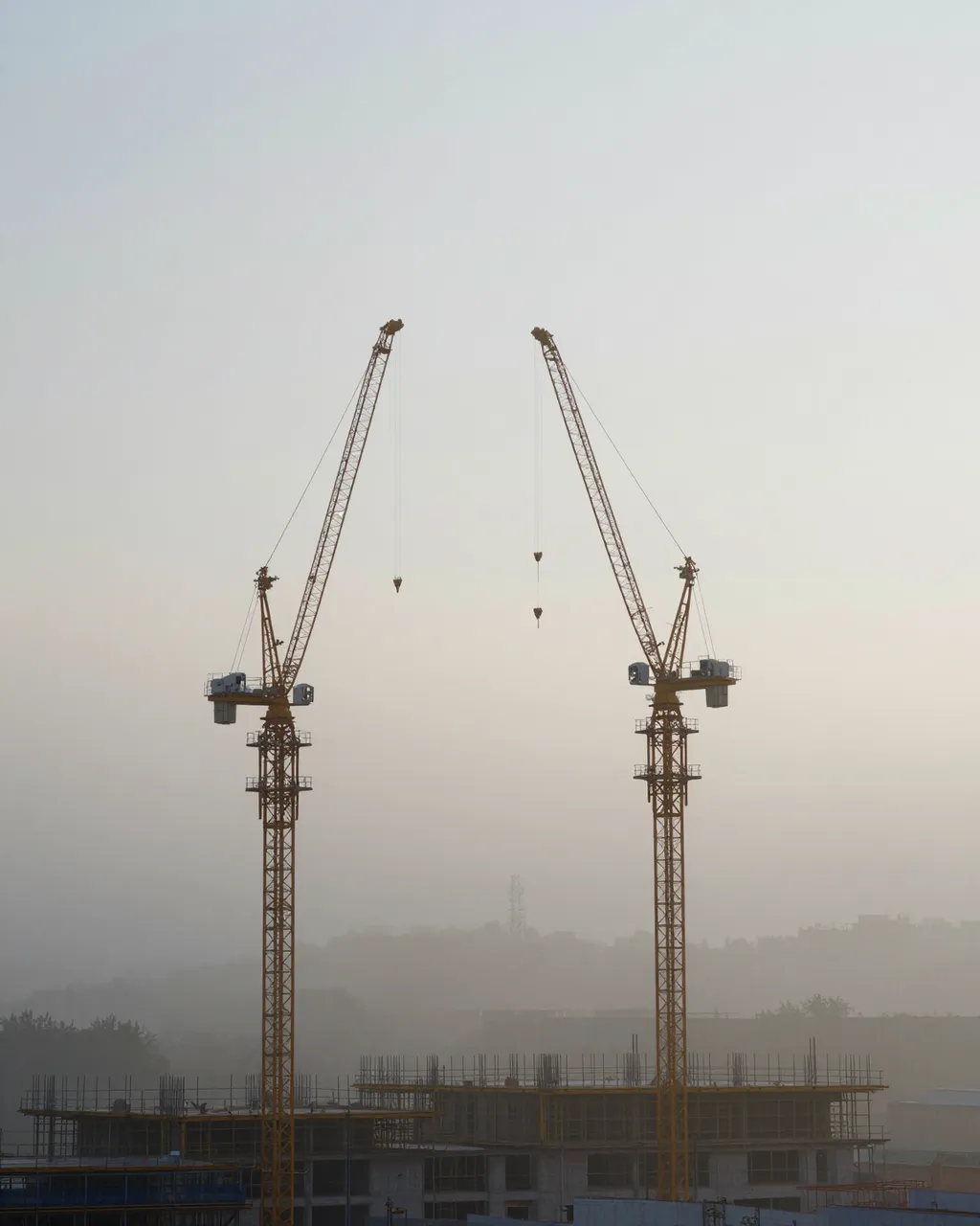 Two tall yellow construction cranes stand over an active building site shrouded in soft morning fog