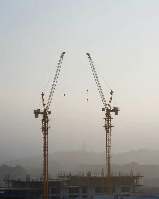 Two tall yellow construction cranes stand over an active building site shrouded in soft morning fog
