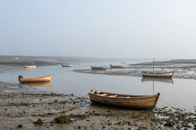 Wooden fishing boats rest on calm still water and exposed mud in a thick foggy small harbor