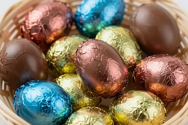 Shiny foil-wrapped and unwrapped chocolate Easter eggs sit inside a light wicker basket.