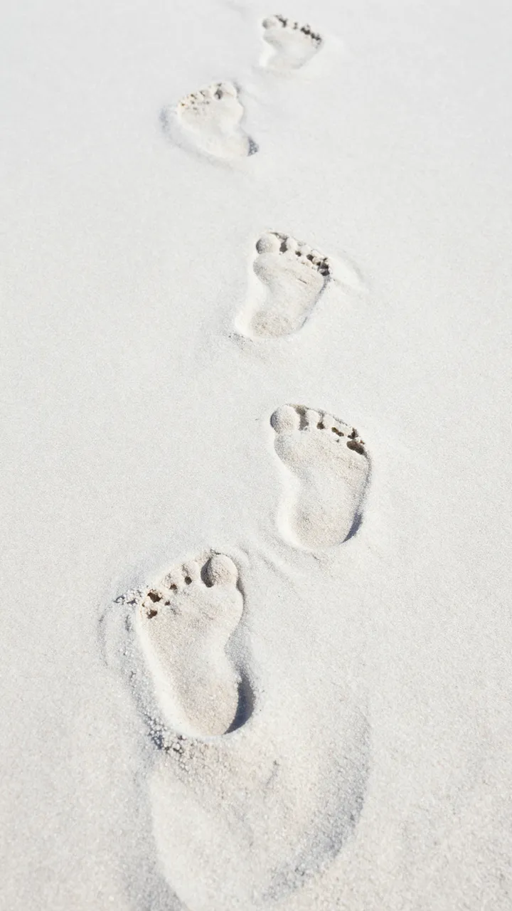 A vertical trail of bare human footprints stretches across fine pale white beach sand.