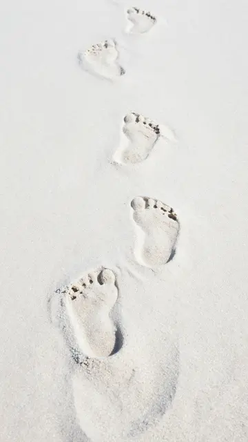 A vertical trail of bare human footprints stretches across fine pale white beach sand.