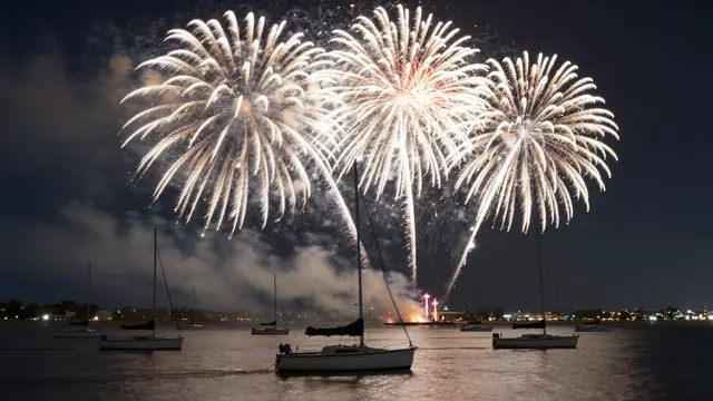 Bright white fireworks burst over anchored sailboats on a calm night harbor