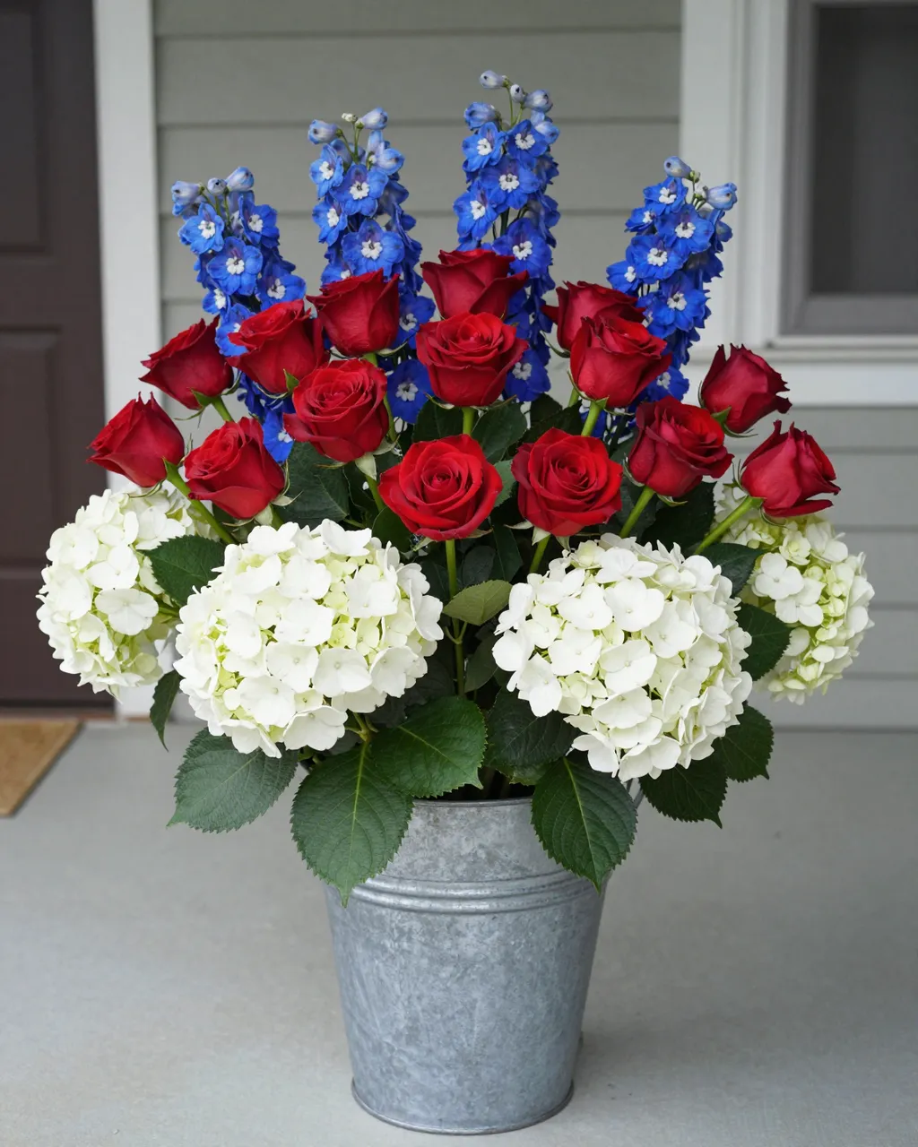 Red roses, white hydrangeas, and blue delphiniums arranged in a galvanized metal pail on a home porch