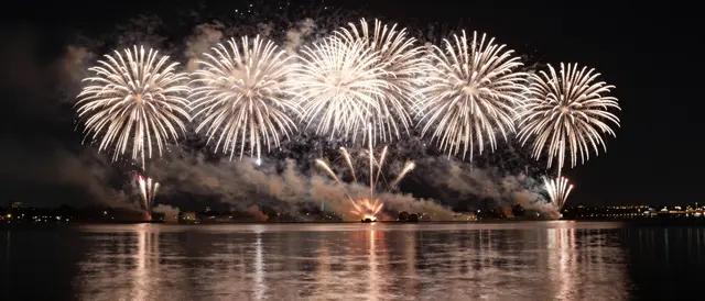 Panoramic golden fireworks burst over calm night water with glowing mirrored reflections